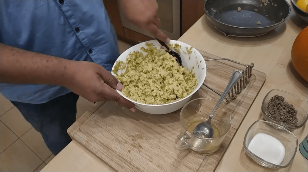 Someone mixes stuffing ingredients including crumbled cornbread, sauteed vegetables, and turkey broth in a large white bowl. A few kitchen utensils and bowls of salt and sage sit on the counter around it.
