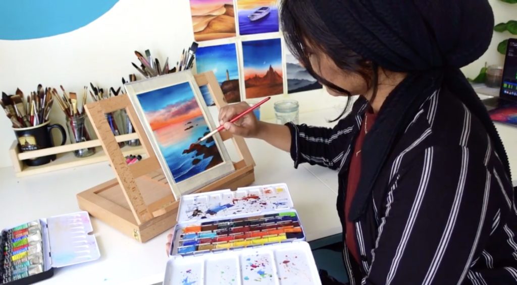 Artist Zaneena Nabeel sits at a desk, holding a paintbrush and a palette of watercolors. She is working on a watercolor scene of the seashore at sunset. In the background, her brushes, water cup, and other paintings are visible.