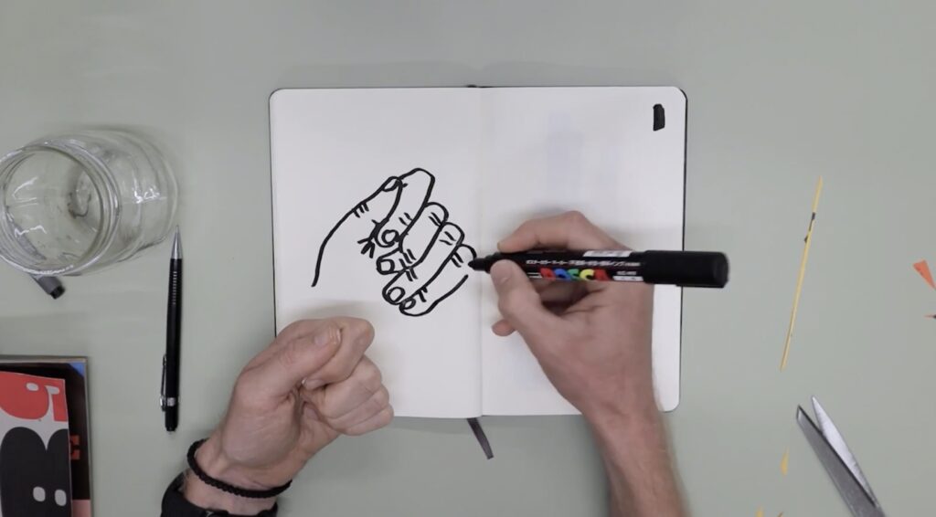 An overhead shot of two hands and a notebook. The right hand is drawing an illustration of the left one in the notebook, using a large black marker.