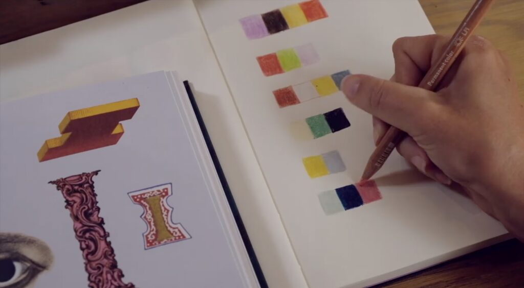 An overhead shot of a notebook. A hand is visible, holding a colored pencil, which is creating one of many colored squares in a palette she’s creating. On the left hand side, some reference images, such as an eye and some stylized letters, are visible. 