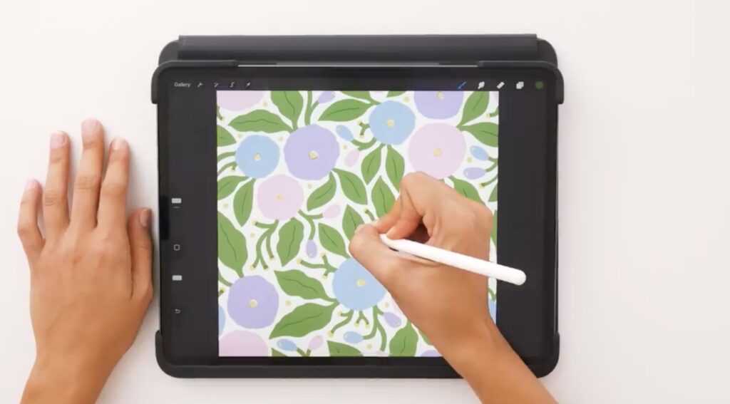 An overhead shot of an iPad displaying a blue, pink and purple floral pattern illustration. Artist Di Ujdi’s hands, one of which is holding an Apple Pencil, are visible in the shot. 