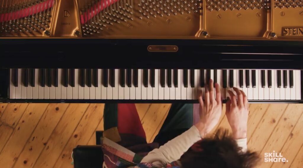 An overhead shot of teacher Jacob Collier playing the piano. His hands are visible on the keys; beneath the piano is a hardwood floor.