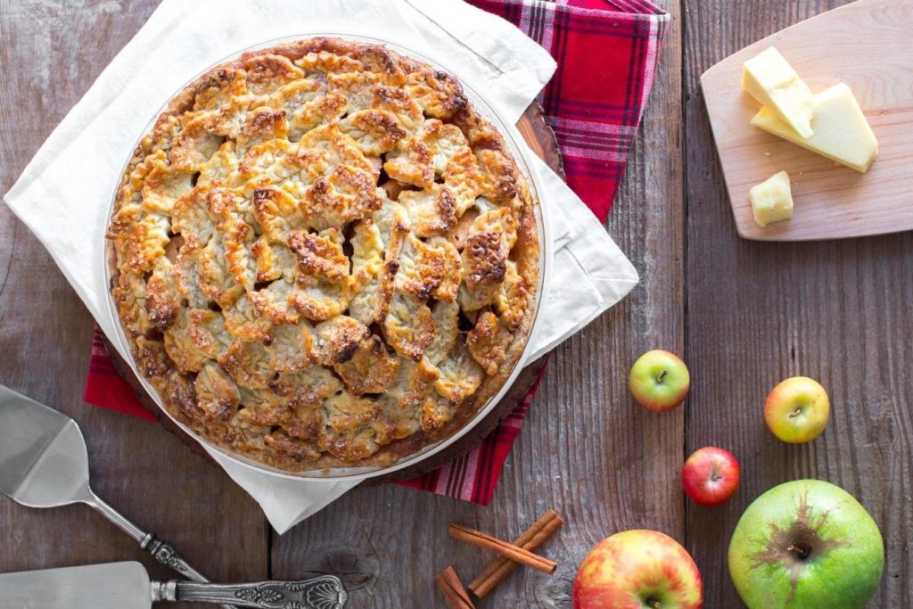 A crispy, sugar crusted apple cheddar pie sits in the top left of the screen. Around its edges are two pie servers, five apples, and a few chunks of cheddar cheese. 