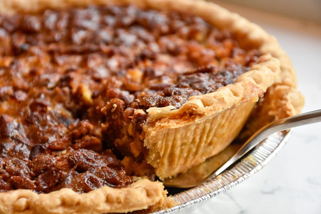 A gooey slice of pecan pie is being taken out a silver pie tin with a pie server. 