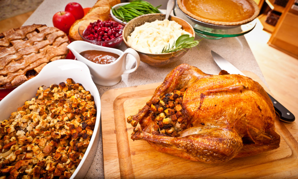 A golden-brown stuffed Thanksgiving turkey sits on a cutting board. Next to it is a dish of stuffing, and behind these two dishes sit pies, a full gravy boat, mashed potatoes, cranberries, and green beans