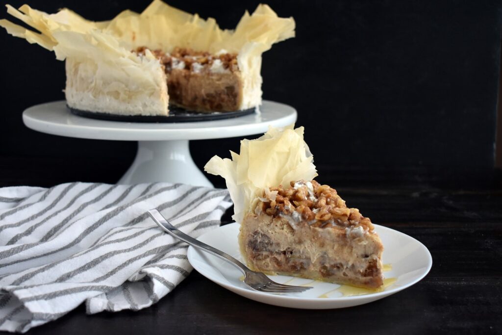 A slice of baklava cheesecake placed in front of an entire cake in the background. You can see the layer of cheesecake filling and chopped walnuts on the top, with the phyllo pastry spilling out the top and side.