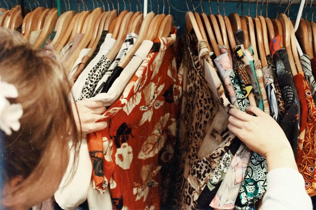 A woman with brown hair flips through a collection of colorful patterned tops on hangers. She is facing away from the camera, so all we see is the back of her head, the side of her cheek, and her hands on the clothing.