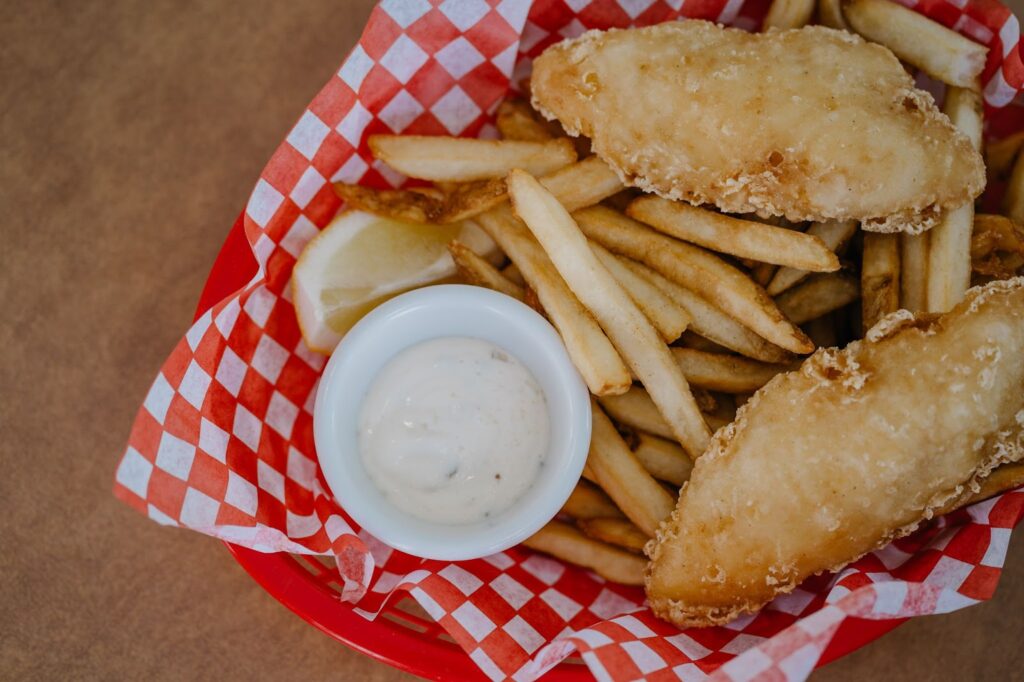 Two fried fish tenders sit in a red plastic basket accompanied by french fries, tartar sauce, and a lemon slice.
