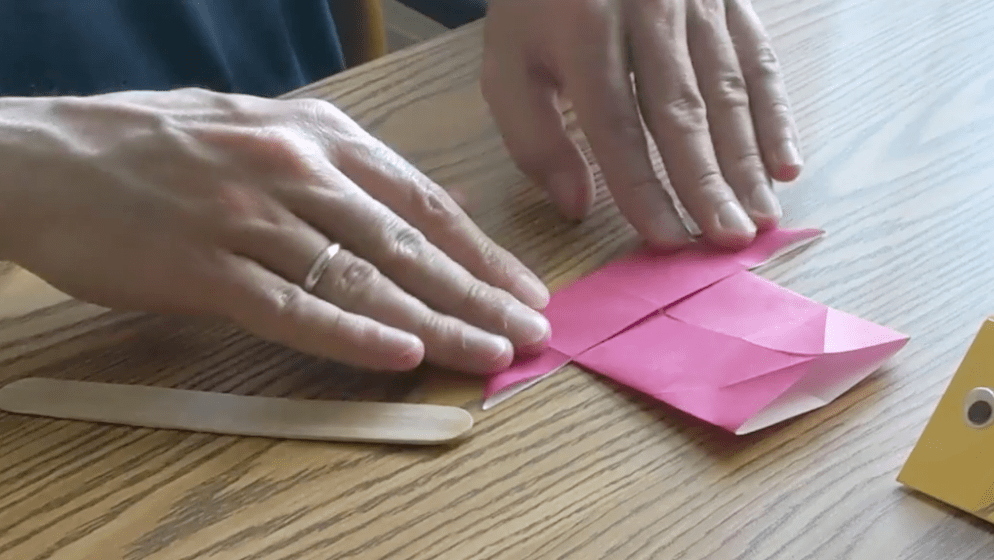 Pink paper is folded into a sailboat shape to start defining the edges of the origami fish.