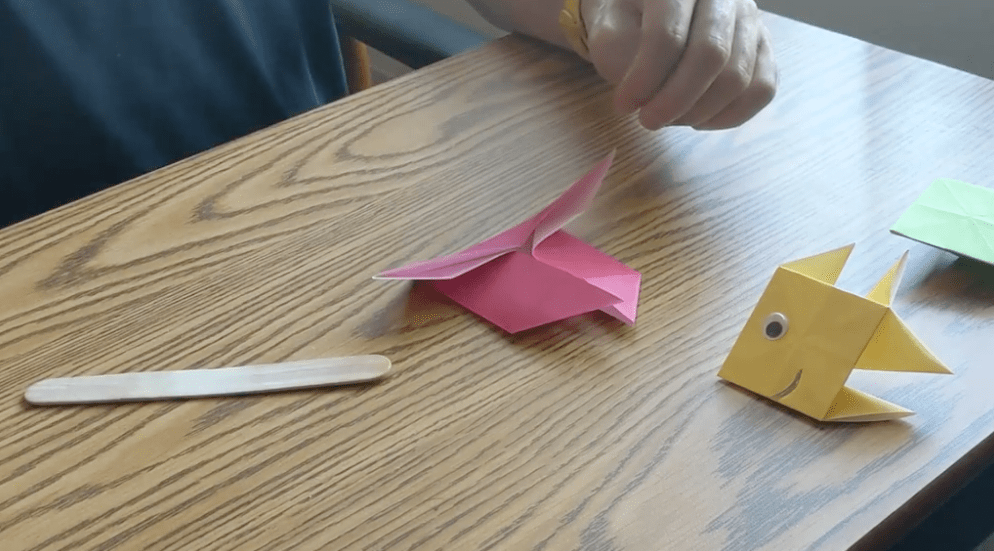A completed yellow origami fish sits on a desk with a stick used to firmly crease the edges of a pink work-in-progress paper fish.