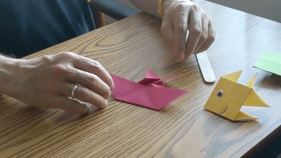 The pink origami paper returns to a sailboat shape before making up the final design of a fish, like the completed yellow one sitting on the table.