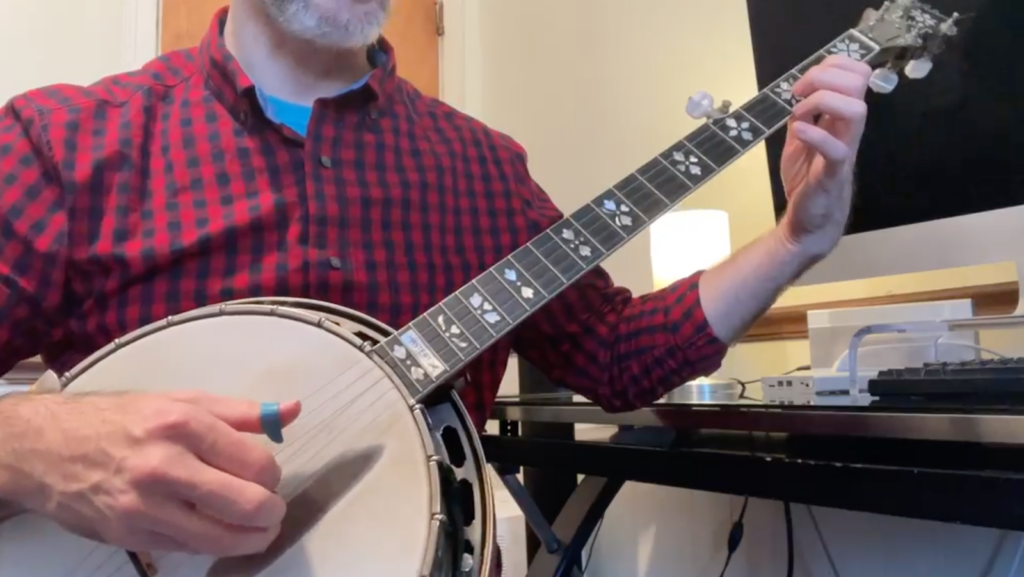 A man in a red and black gingham shirt plays the banjo. His left hand holds the banjo by its neck and his right hand has three fingers on its strings.