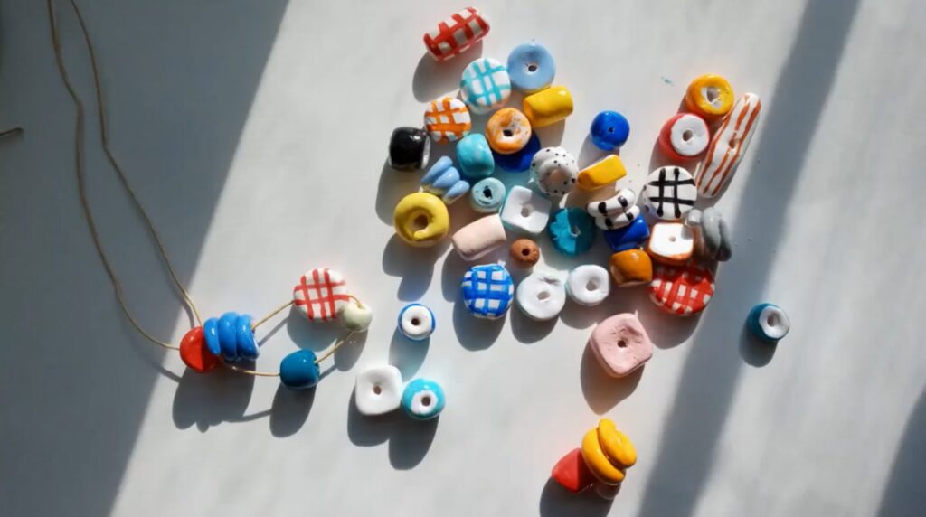 An assortment of colorful clay beads lays in a sunbeam on a white tabletop. A few of the beads are strung on a necklace, while the rest are loose.