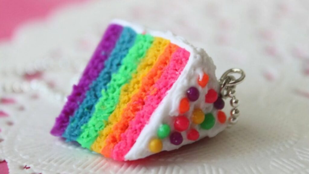 A close-up of a tiny charm made from polymer clay. The clay has been sculpted into the shape of a slice of rainbow cake, and is decorated with white frosting and vibrant sprinkles.