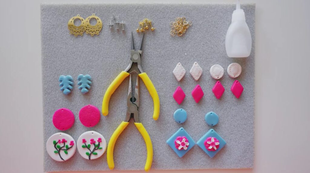An assortment of white, hot pink and light blue polymer clay earrings arranged on a gray felt mat, along with two pairs of pliers and a small bottle of glue.