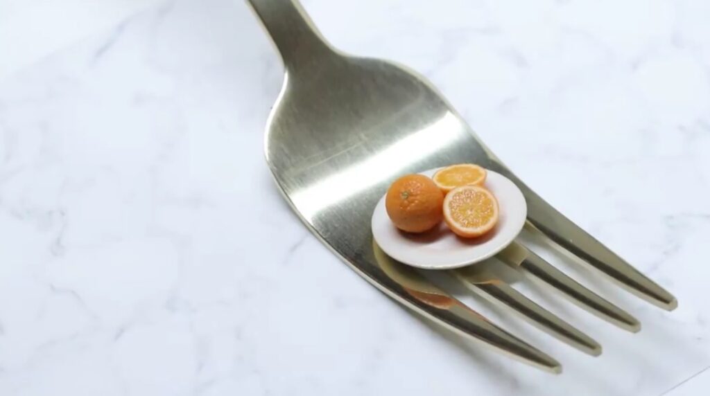 A silver for resting on a white granite countertop. Resting on the middle of the fork is a tiny white plate of miniature orange slices and one intact orange.