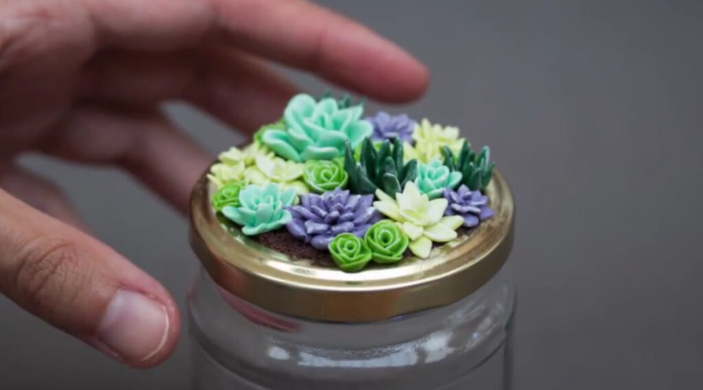 A woman’s hand next to a glass jar with a gold lid. On top of the lid is a tiny succulent garden, with each of the plants being crafted from polymer clay.