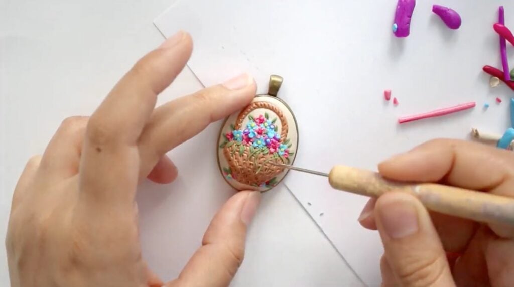 A woman’s left hand holding an oval pendant decorated with a basket of flowers sculpted from polymer clay. With her right hand, she uses a metal implement to add blades of grass to the basket.