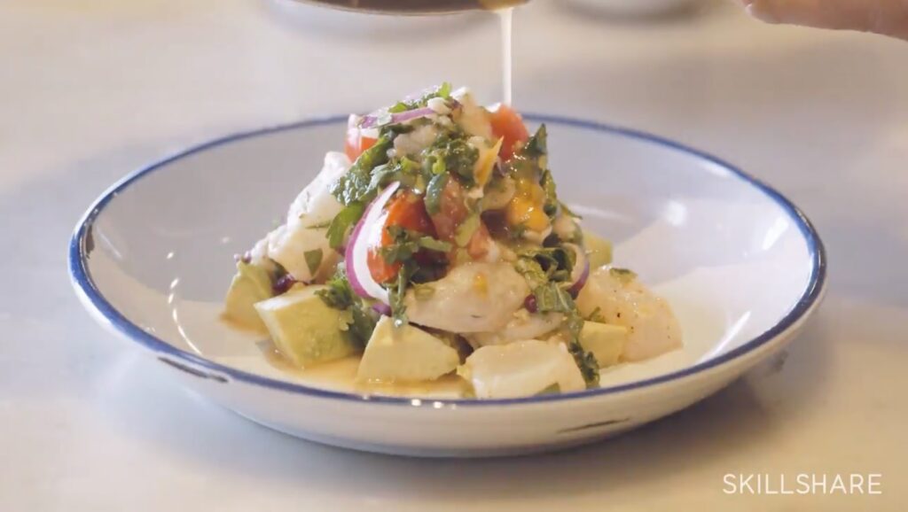 In a white dish with a blue rim, a serving of ceviche lays on top of a few chunks of avocado. Its citrusy sauce is being poured over the onion, tomato, raw fish, and herb combination.