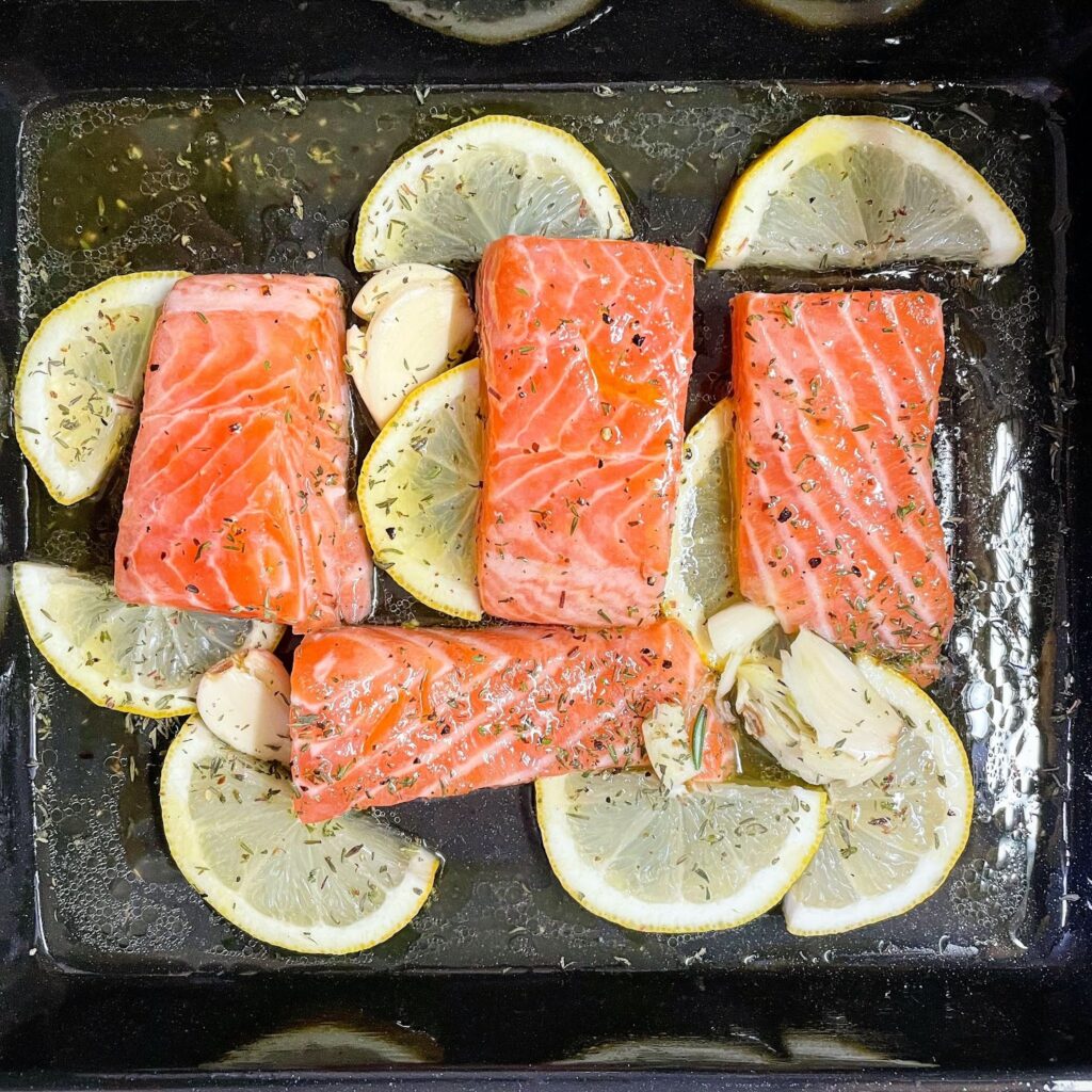 Four salmon filets sit in a pan surrounded by smashed garlic, olive oil, and lemon slices.