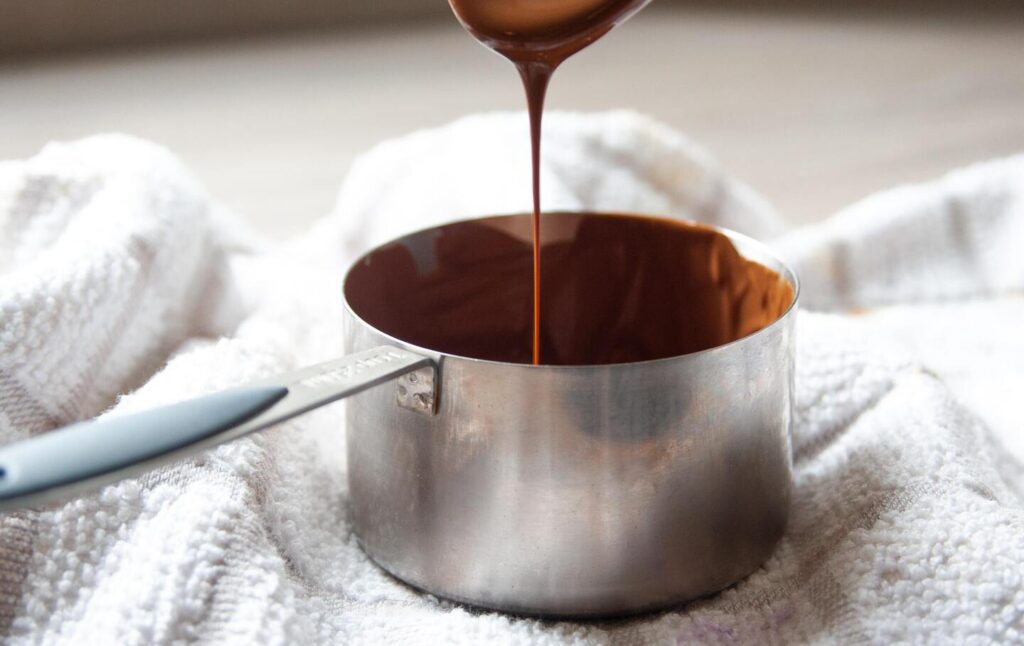 A saucepan is placed on top of a dishcloth while melted chocolate is poured into it from above.