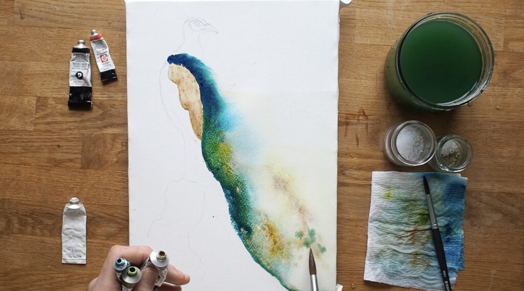 A watercolor painting of a peacock mid-process. The sketched bird is faintly visible on the paper, and a large swatch of mixed watercolor in blues and greens stretches out across the page. A few tubes of paint, a paintbrush, a glass of paint water, and one of Davies’ hands are also visible in the shot.