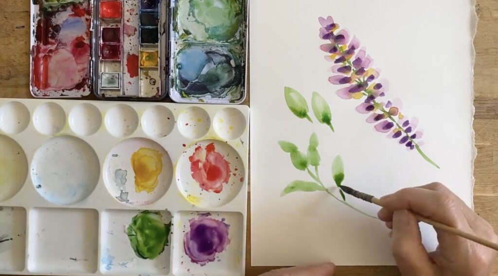 A mid-process watercolor painting of lavender sprigs. Also in shot is a watercolor palette, a tray of gouache paint, and Cook’s hand, painting a green leaf.