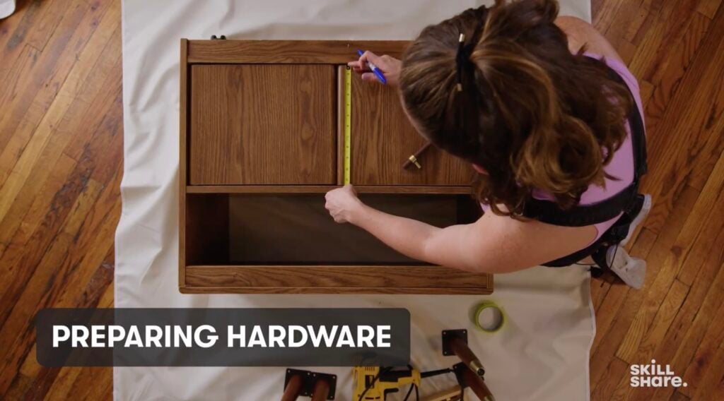 An overhead shot of Tarboton measuring the width of a wooden cabinet. The cabinet is sitting on a white dropcloth, on top of a hardwood floor.