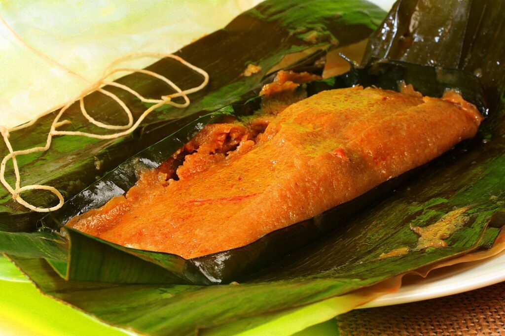 An unwrapped pastel sits within a banana leaf and some parchment paper. Unwrapped butcher’s twine is visible in the back corner.