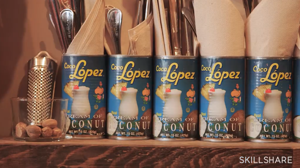 Cans of creamed coconut sitting on a wooden shelf in a bar, alongside cocktail garnishes like nutmeg and a grater.