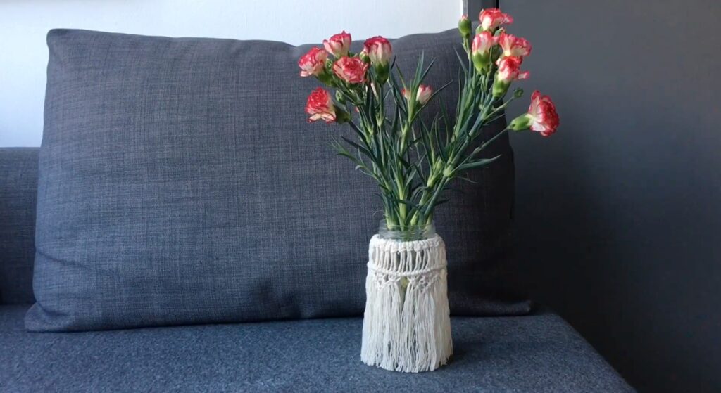 A bunch of pink carnations are sat in a glass jar that’s hidden behind a macramé textile. The top third is made of knots, while the bottom two thirds is made of fringing that hangs to the bottom. The vase is placed on a gray couch.