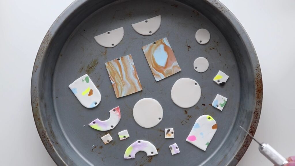 Several unbaked polymer clay earring pieces in plain, marble and terrazzo patterns are arranged on a baking pan and ready to go into the oven.