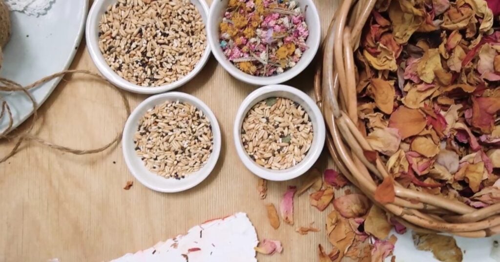 Looking down at a wooden table top full with four small bowls of seeds and dried plant materials next to a large woven basket full of dried flower petals.