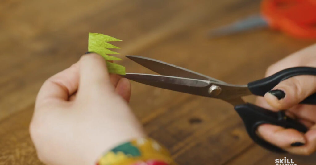 Close up of an artist cutting long triangles into a small, green crepe paper square to look like a patch of grass.
