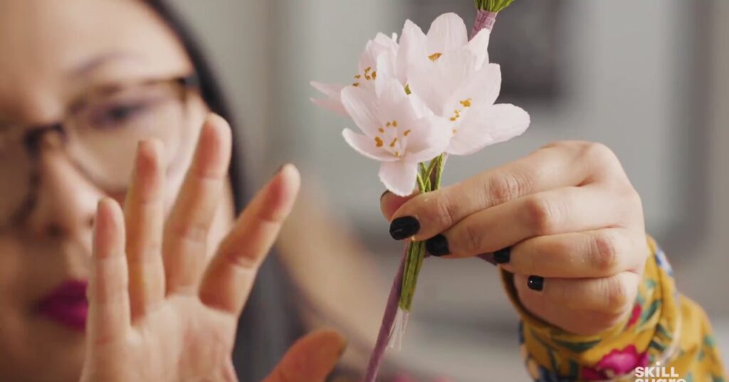An artist holds five crepe paper cherry blossoms against a faux branch, preparing to attach the stems with more paper.