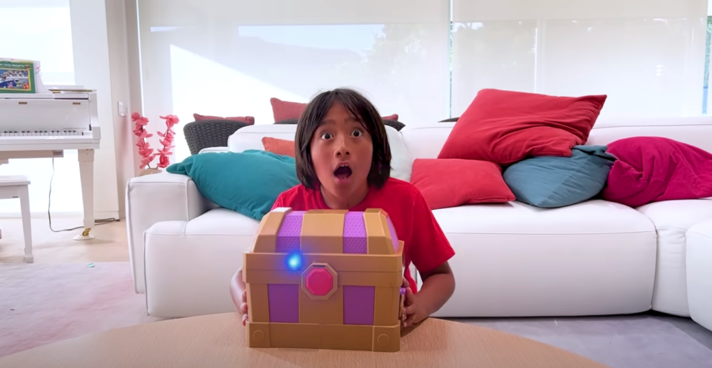 YouTuber Ryan Kaji with a gold and purple treasure chest toy, in front of a white couch with blue and pink throw pillows.