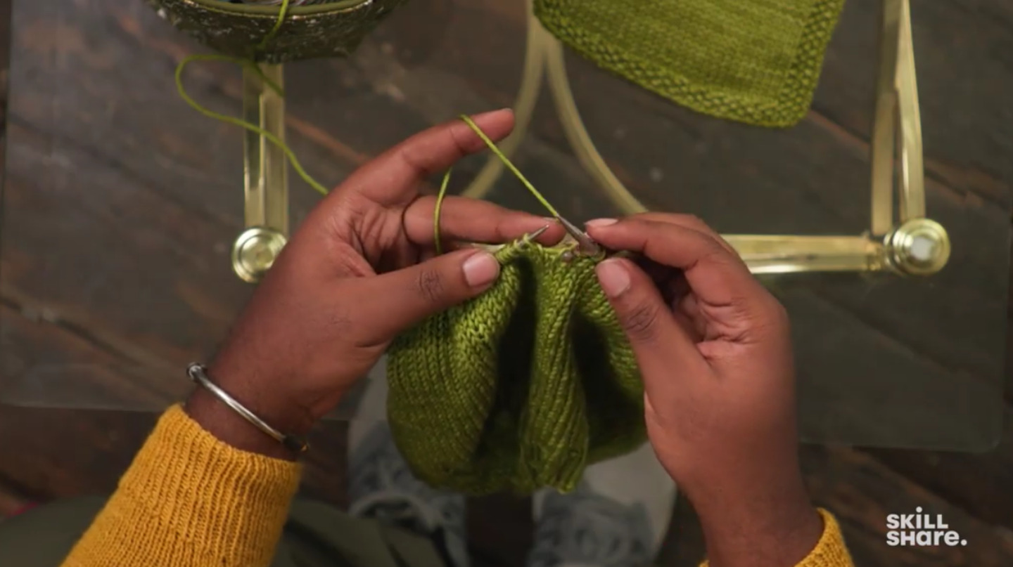 An overhead view of a man’s hands knitting a green cowl with metal knitting needles.