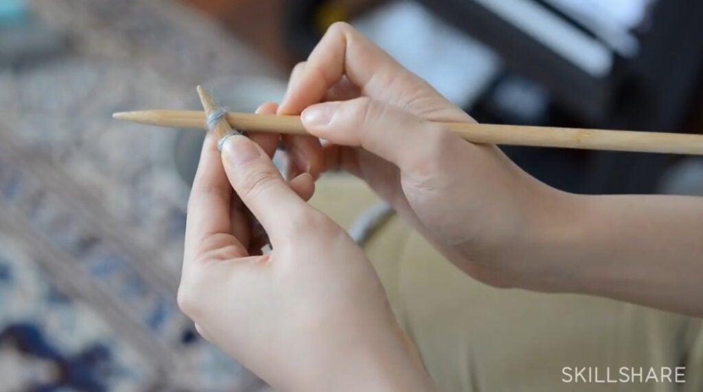 A woman’s hands holding two knitting needles. She’s inserting the right needle into the first stitch on the left needle.
