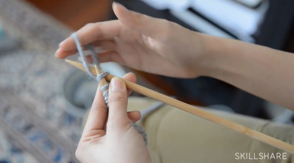 A woman’s hand holding two knitting needles. With her right hand, she’s wrapping the yarn around the tip of the right needle.