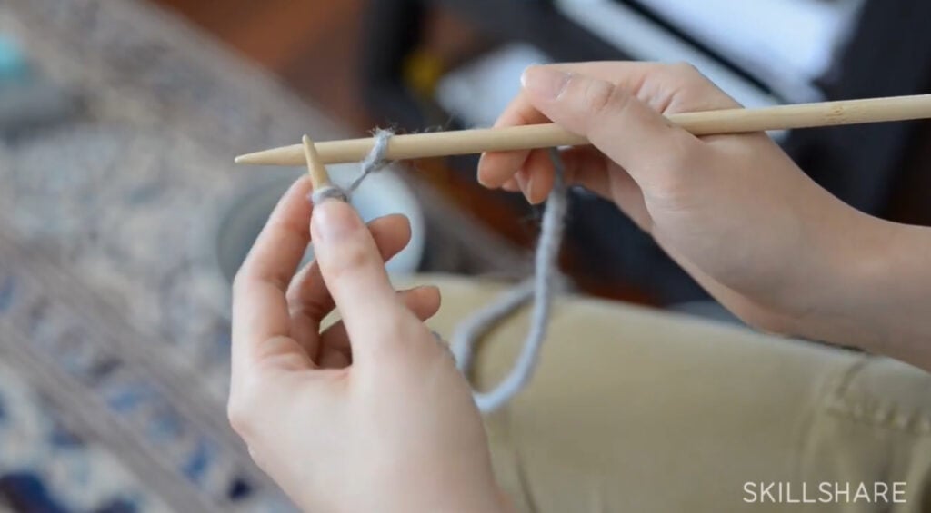 A woman’s hands holding two knitting needles. There’s one stitch on the right needle, which she’s just lifted off the left needle.