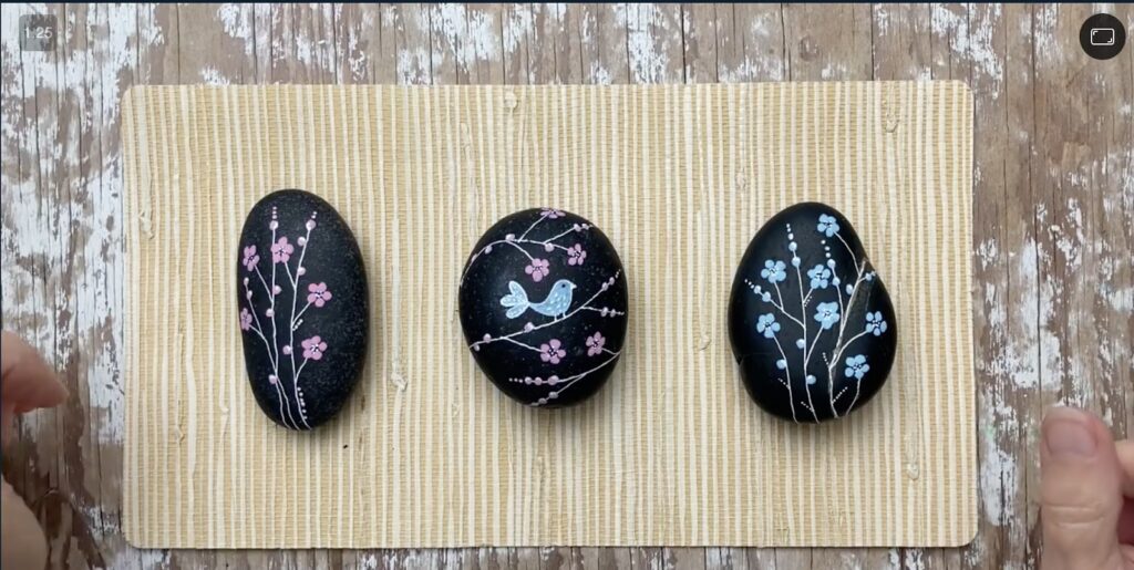 Three black painted rocks sit on a tan mat. They have little flower designs painted on them.