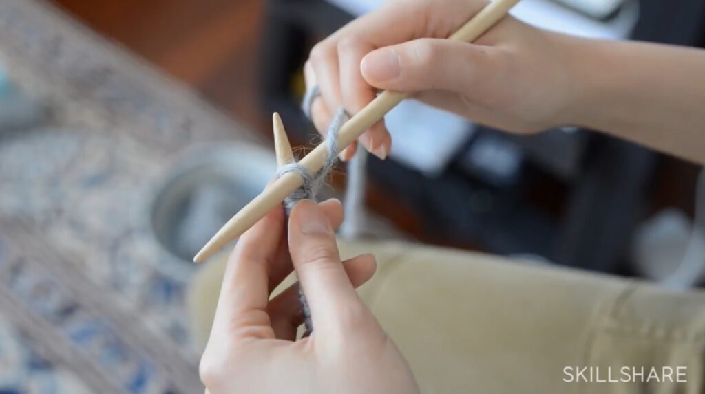 A woman’s hands holding two knitting needles. She’s inserting the tip of the right needle into the first stitch on the left needle, this time from back to front.