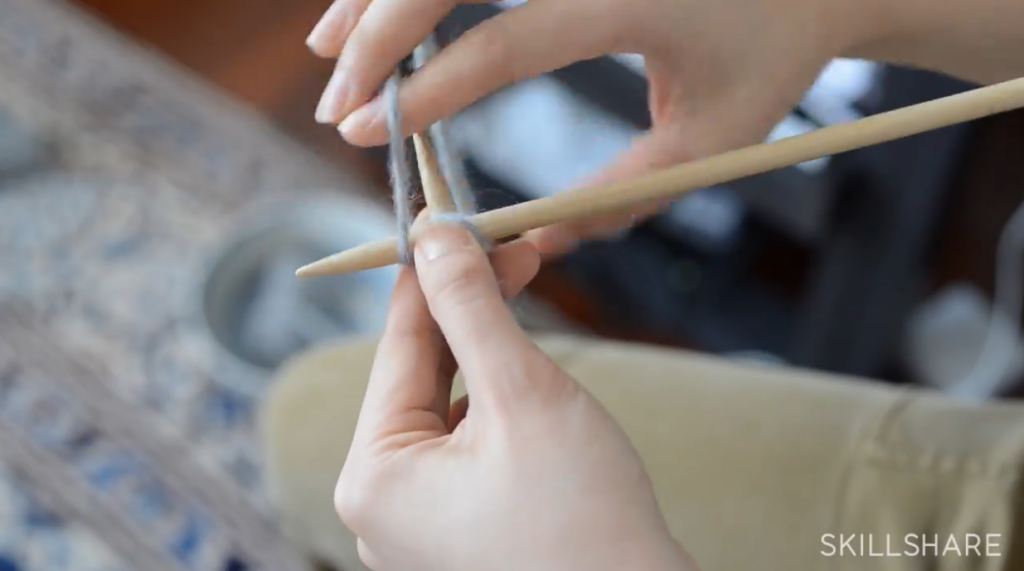 A woman’s hands holding two knitting needles. With the right hand, she wraps the working yarn around the tip of the right needle from back to front and all the way around.