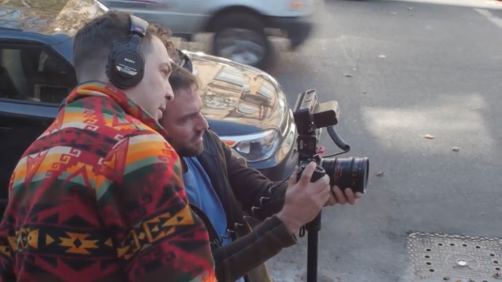 A film director looking into the camera monitor while on set during a street-based scene.