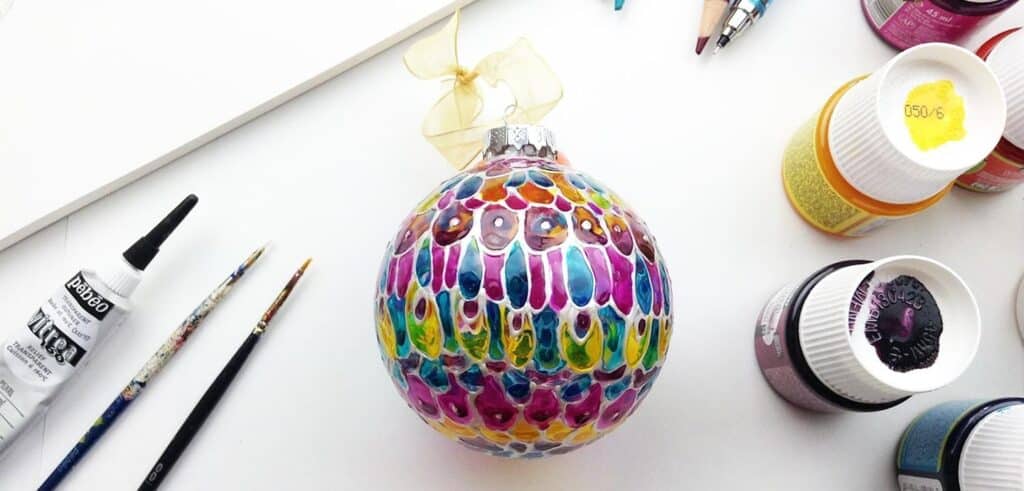 A painted glass ornament with oval and circle patterns in a range of colors, surrounded by the kit you’d need to paint it: glass paint, paint brushes and an outliner.