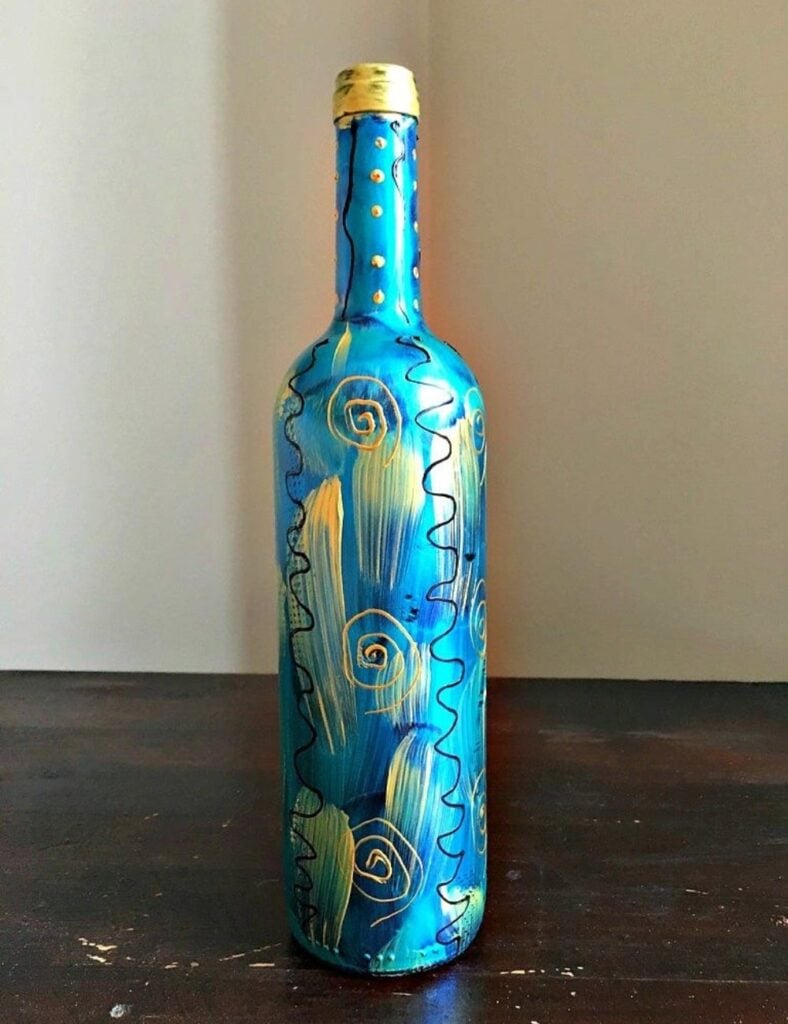 A painted wine bottle with a blue background that’s been decorated with gold and black lines and swirls.