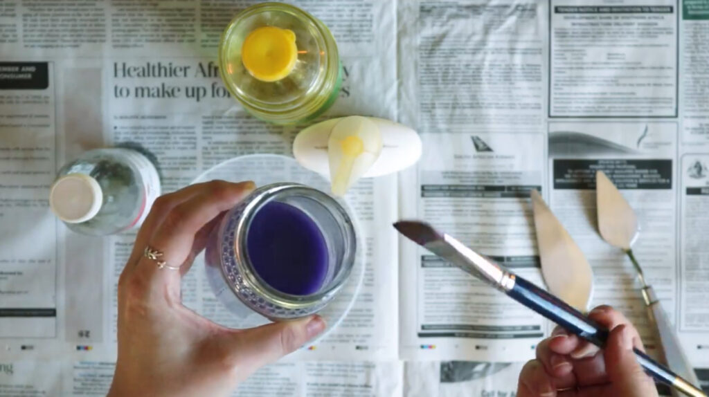 A person's hands holding a paintbrush in one hand and a glass jar full of turpentine in the other.