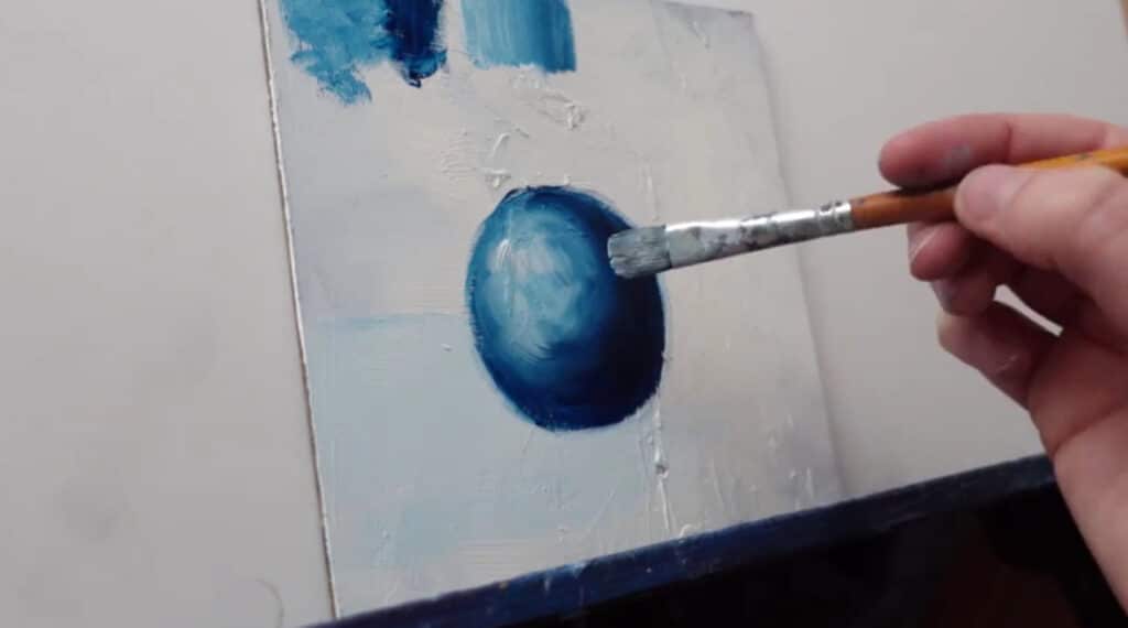 A person's hand painting a small white canvas with an image of a dark blue sphere highlighted with white.