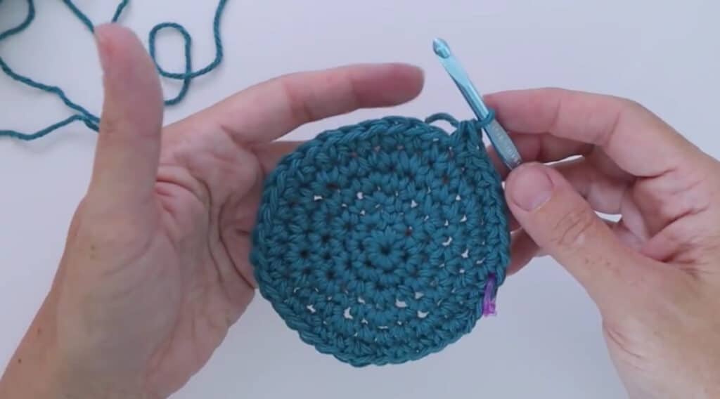 An overhead view of a woman’s hand crocheting a round, dark teal coaster using a blue metal crochet hook.