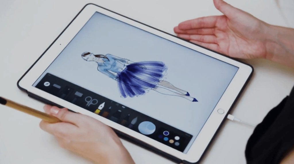 A person's hands resting on either side of an iPad. The iPad screen displays a digital illustration of a woman wearing a light blue button-down shirt and a dark blue pleated tulle skirt.